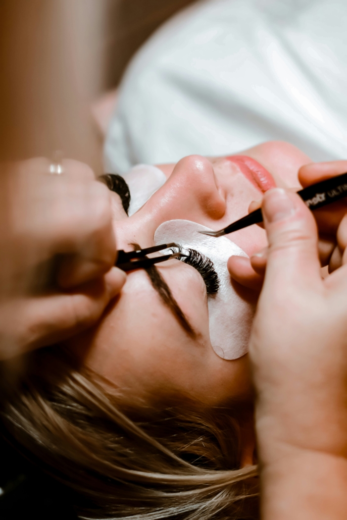 Frau liegt mit Augenpads auf der Liege, während eine Stylistin mit zwei Pinzetten einzelne Wimpernextensions anbringt.